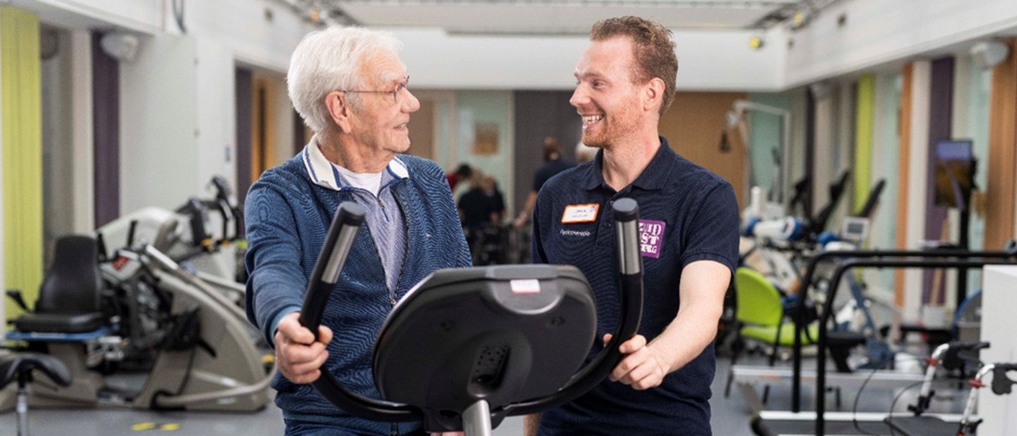 oudere heer bezig op fitnessapparaat, fysiotherapeut aan zijde